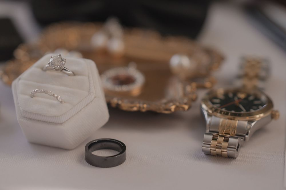 Wedding rings, diamond engagement ring in white velvet box, black tungsten band, and two-tone Rolex watch on mirrored tray.