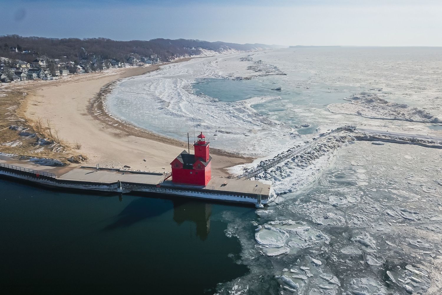 Aerial view of a red lighthouse on a snowy pier surrounded by icy Lake Michigan waters in winter.