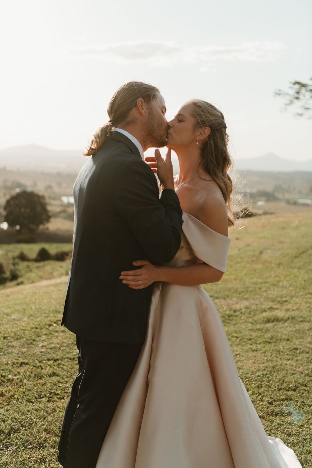 A bride and groom share a romantic kiss at golden hour on a lush green hillside with mountain views.