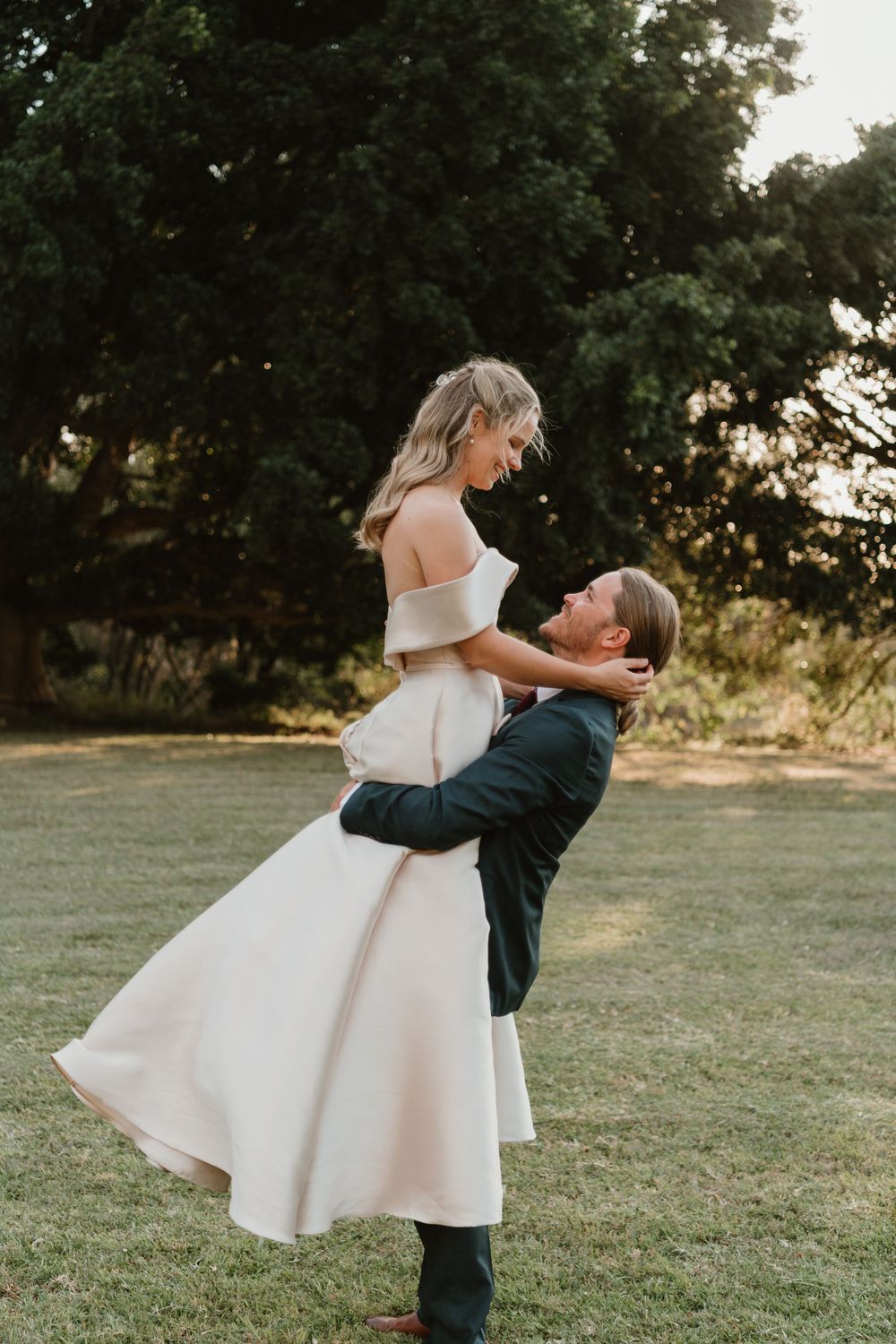 Groom lifting bride in white off-shoulder dress outdoors, sharing joyful moment on green lawn with trees.