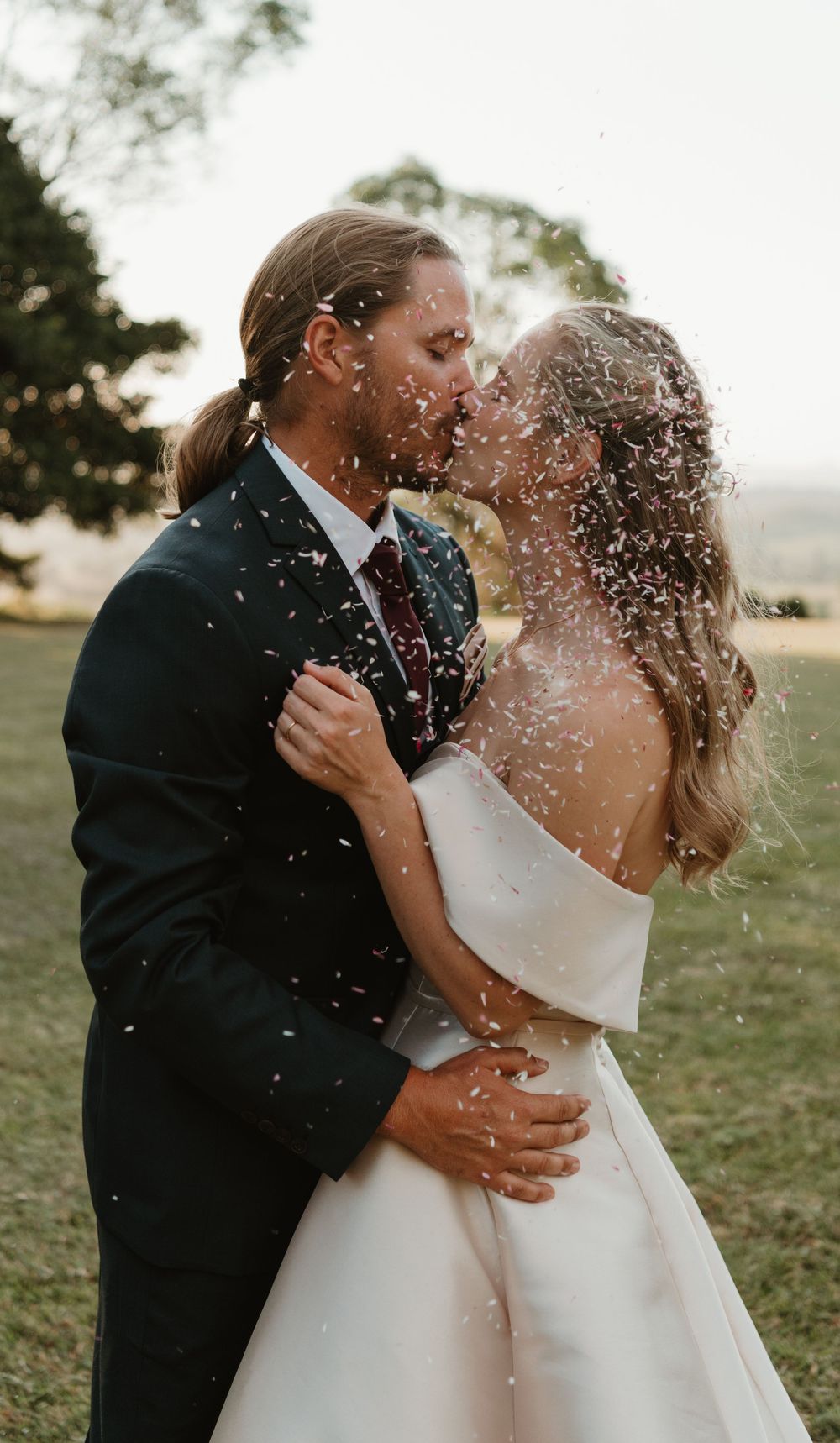 Bride and groom sharing a romantic kiss surrounded by confetti on their wedding day outdoors.