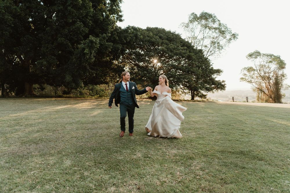 Bride and groom walking hand in hand across lush green lawn with large trees at outdoor wedding venue.