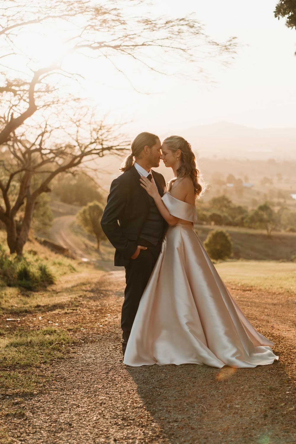 Bride and groom share a romantic moment at golden hour on a scenic gravel path, bride in elegant off-shoulder gown.