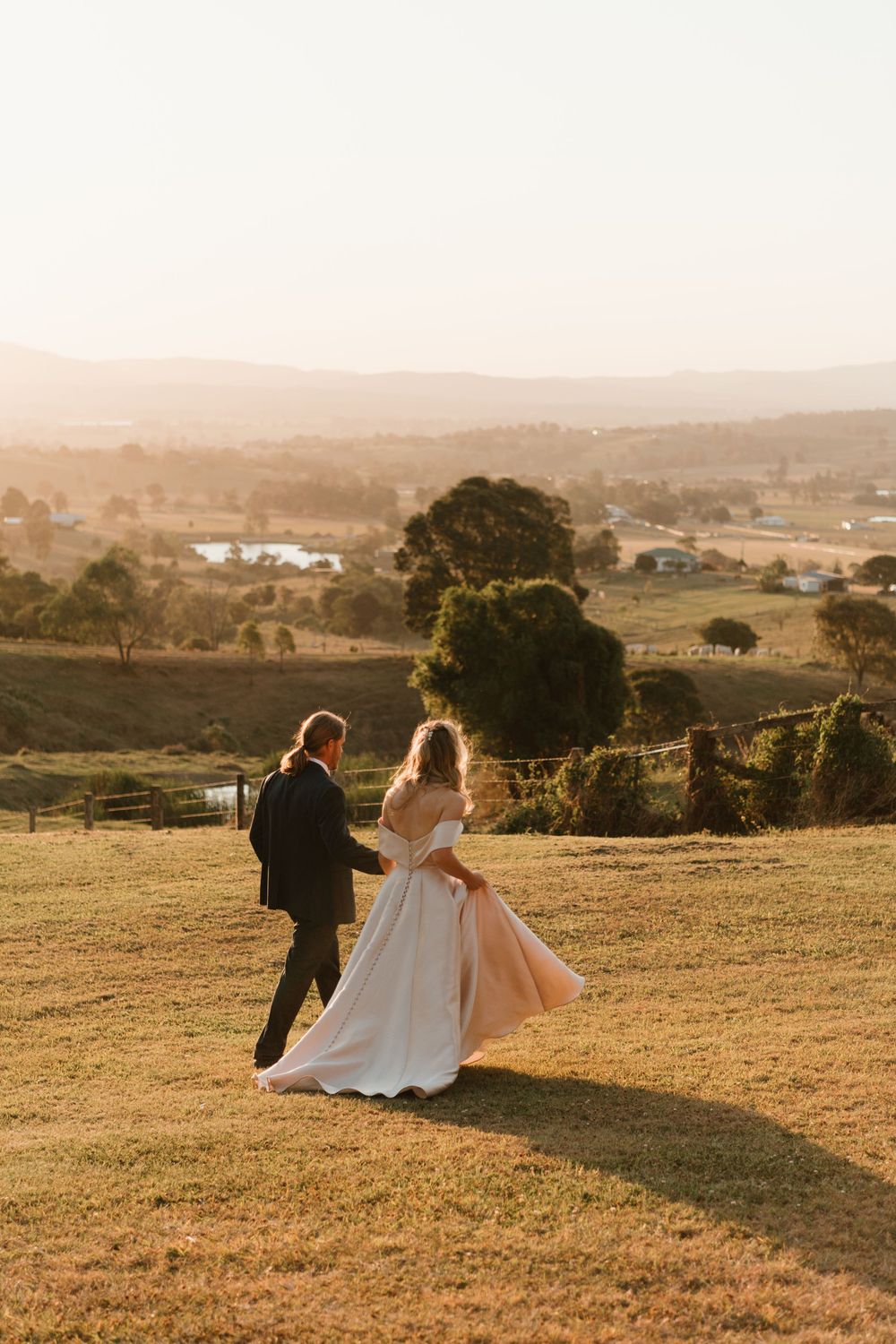 Bride and groom walk hand in hand across golden sunset hillside overlooking scenic valley landscape.