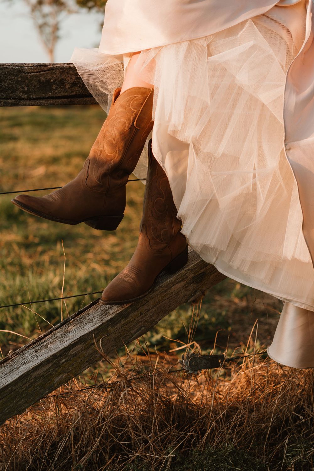 Woman in a blush tulle skirt and brown cowboy boots sitting on a rustic wooden fence at golden hour.
