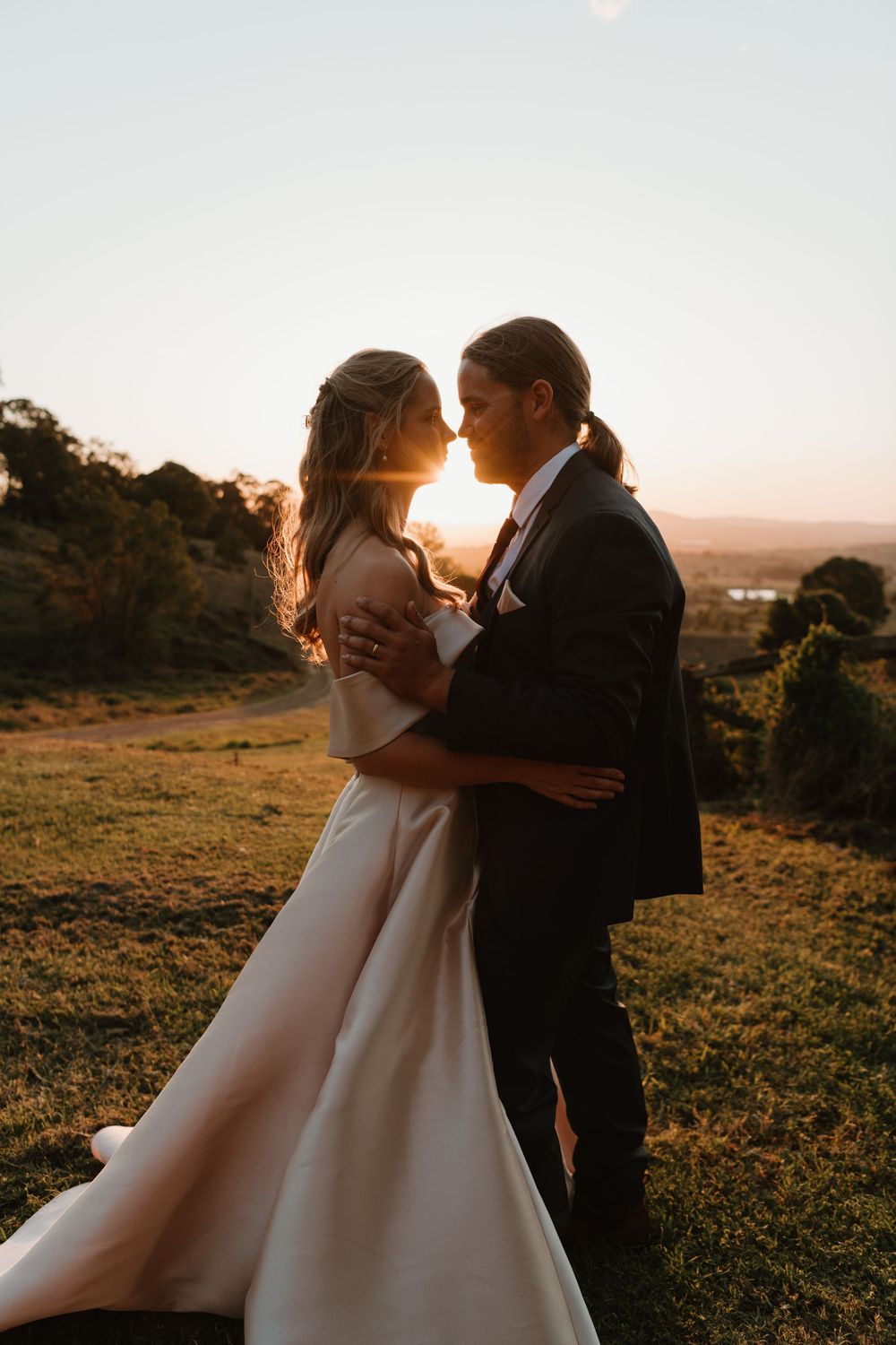 A bride and groom share a romantic sunset embrace outdoors, with golden backlight illuminating their wedding attire.