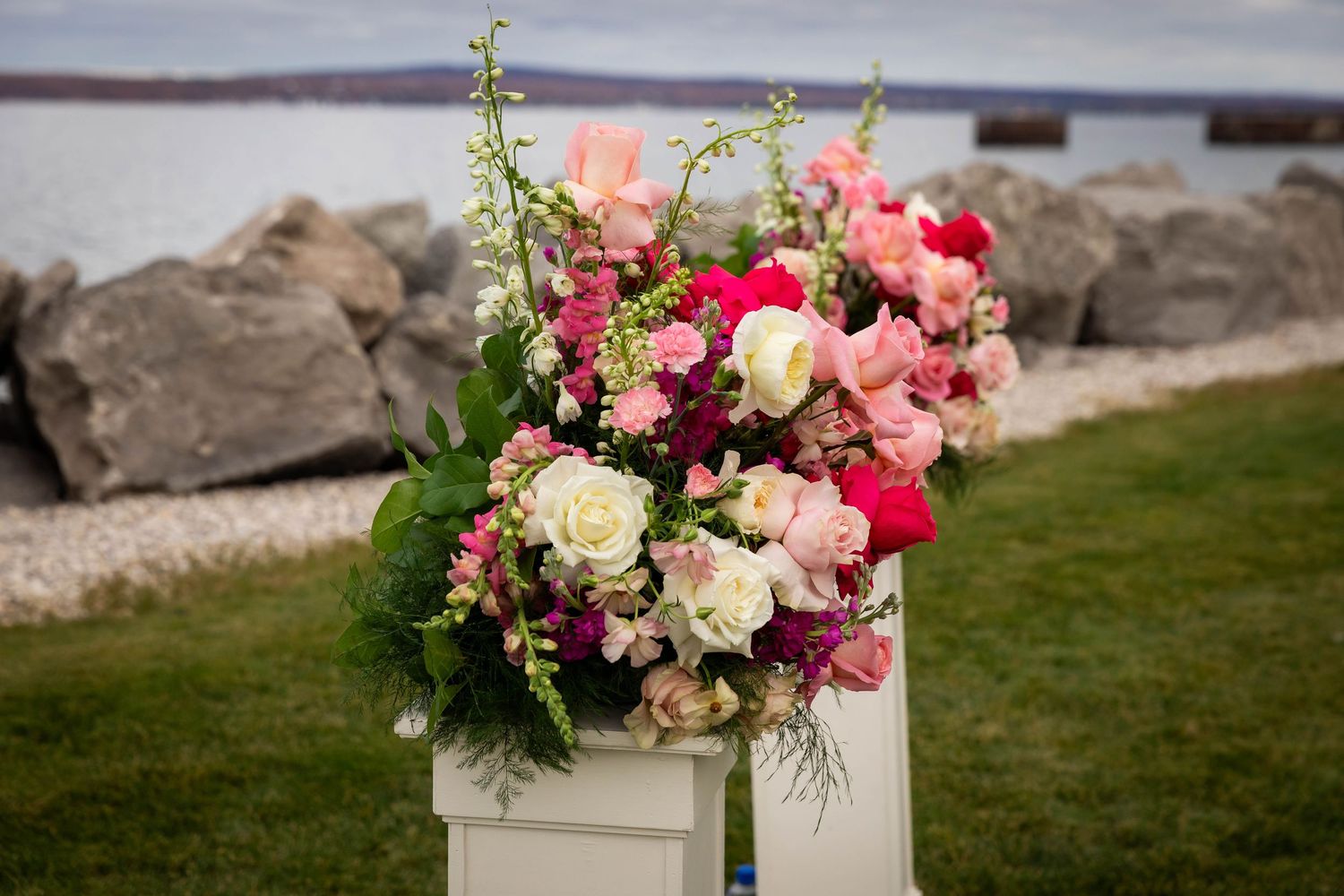 Elegant floral arrangement of pink, red, and white roses on a white pedestal by a rocky waterfront.
