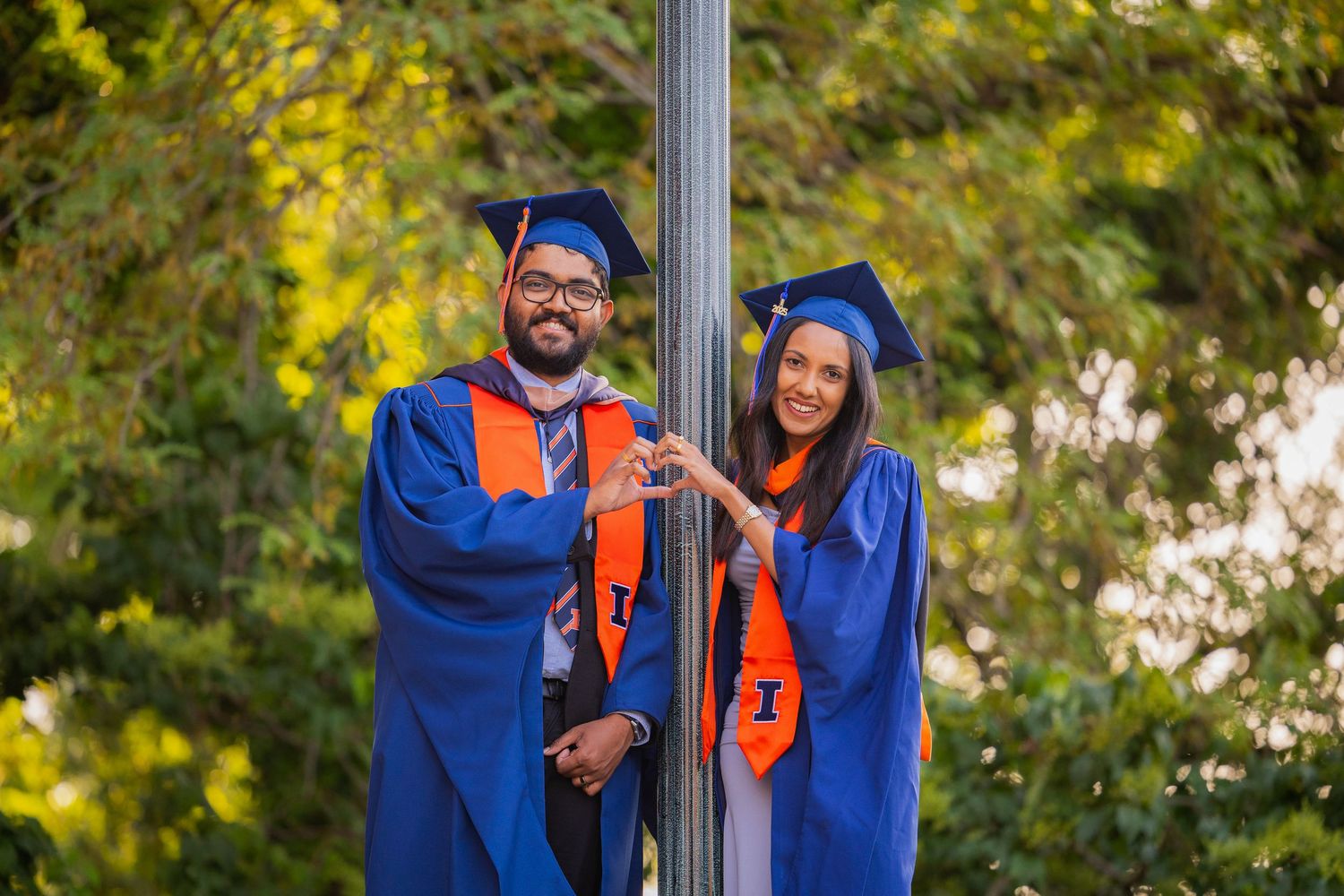 Couple graduating standing beside each other leaning against a lamp post doing heart hands at UIUC