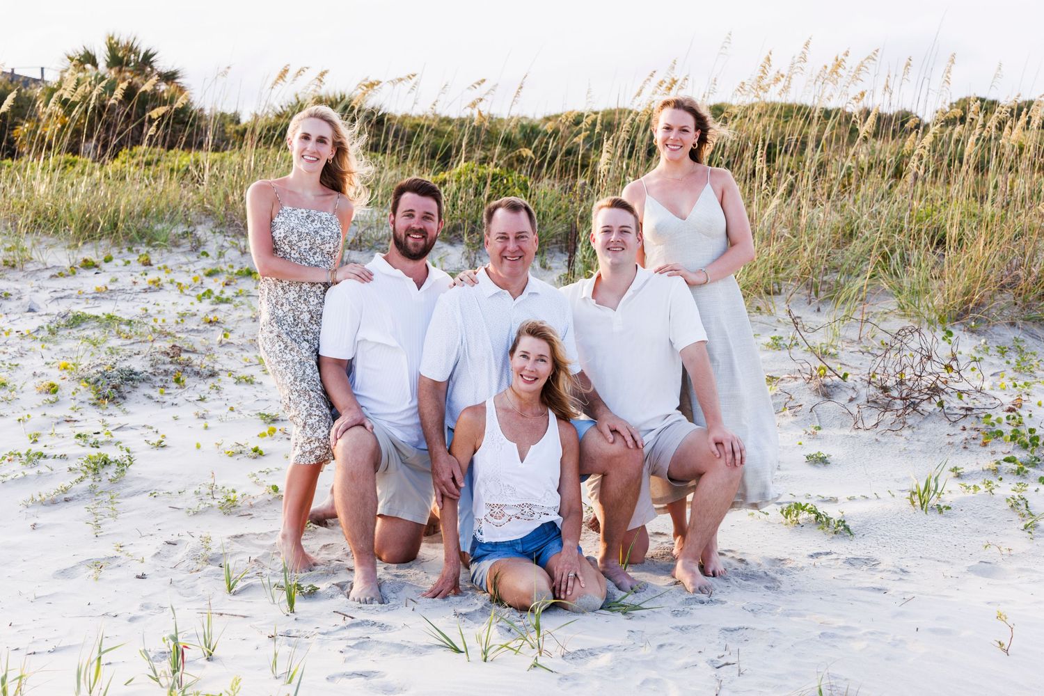 A group sits together on a sandy beach wearing light colored clothing during a photo session at sunset.
