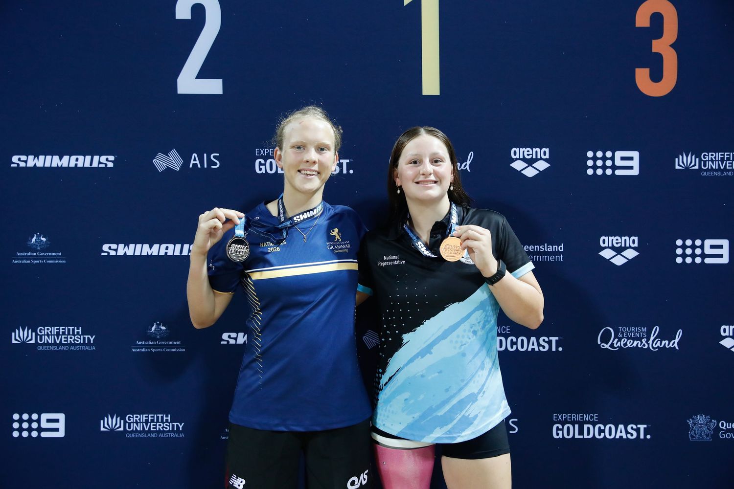 Three female swimmers pose on podium holding medals at Griffith University swimming competition in Queensland, Australia.