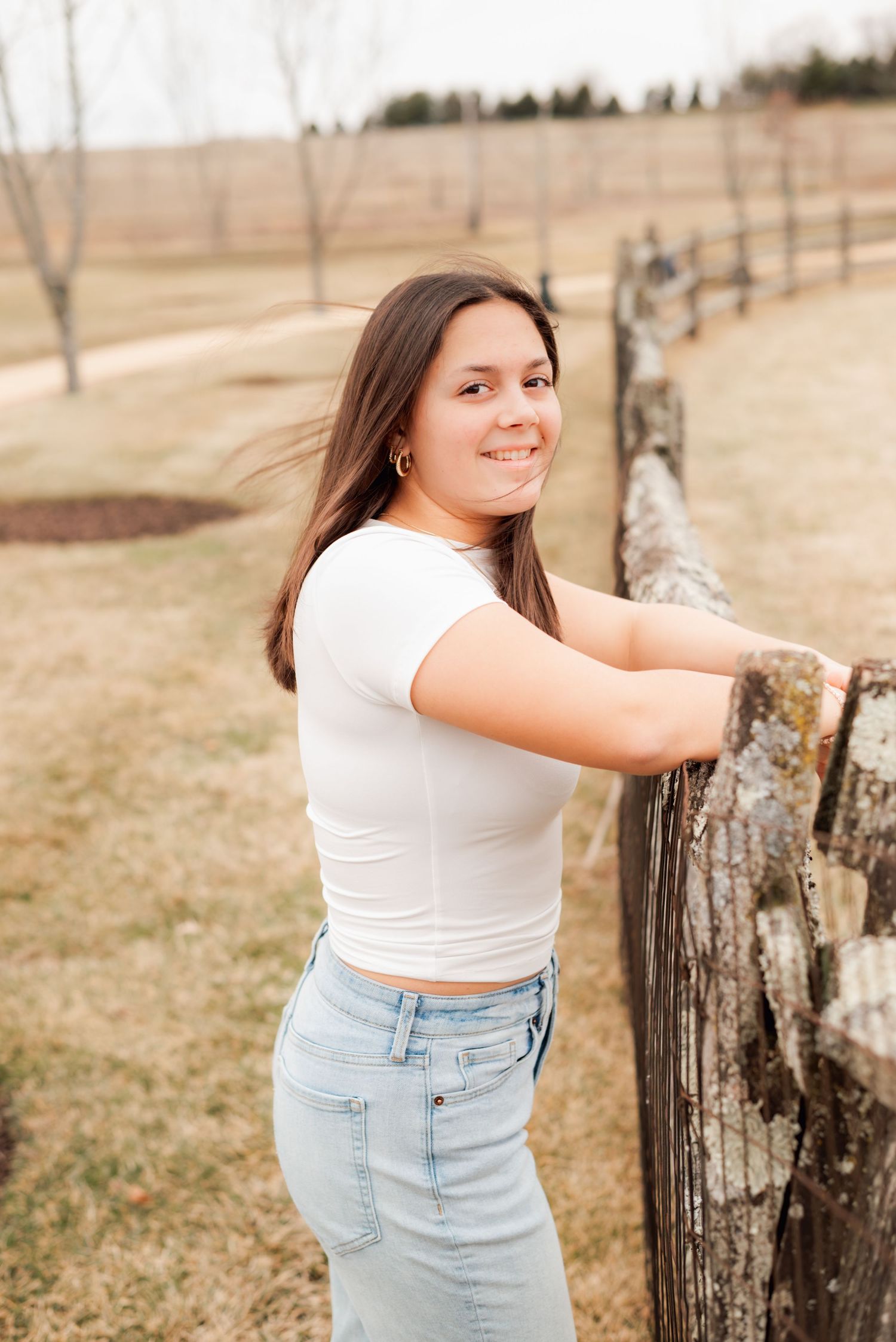 Young woman in white crop top and jeans smiling by a rustic wooden fence in a scenic outdoor setting.
