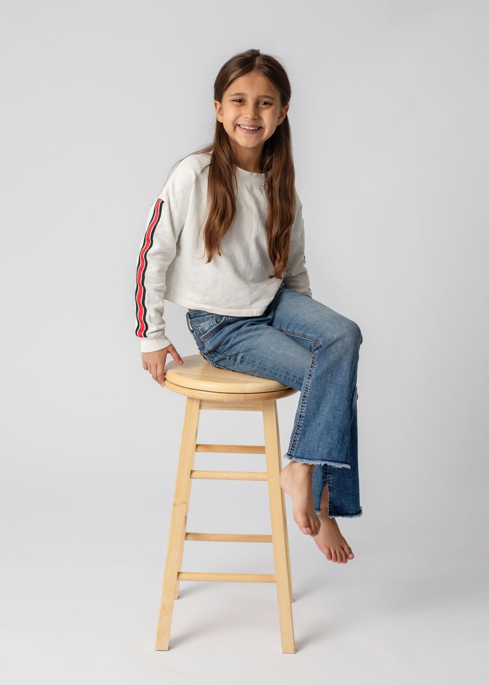 Person in white sweater and blue jeans sitting casually on wooden stool against light gray background.