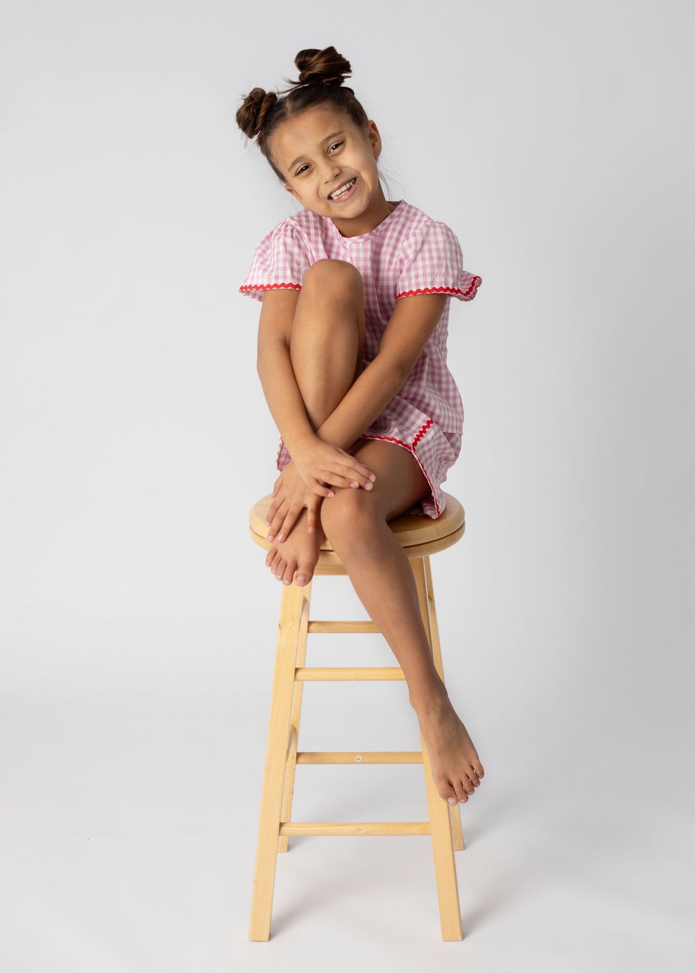 Young child in pink gingham dress sits playfully on wooden stool against gray background.