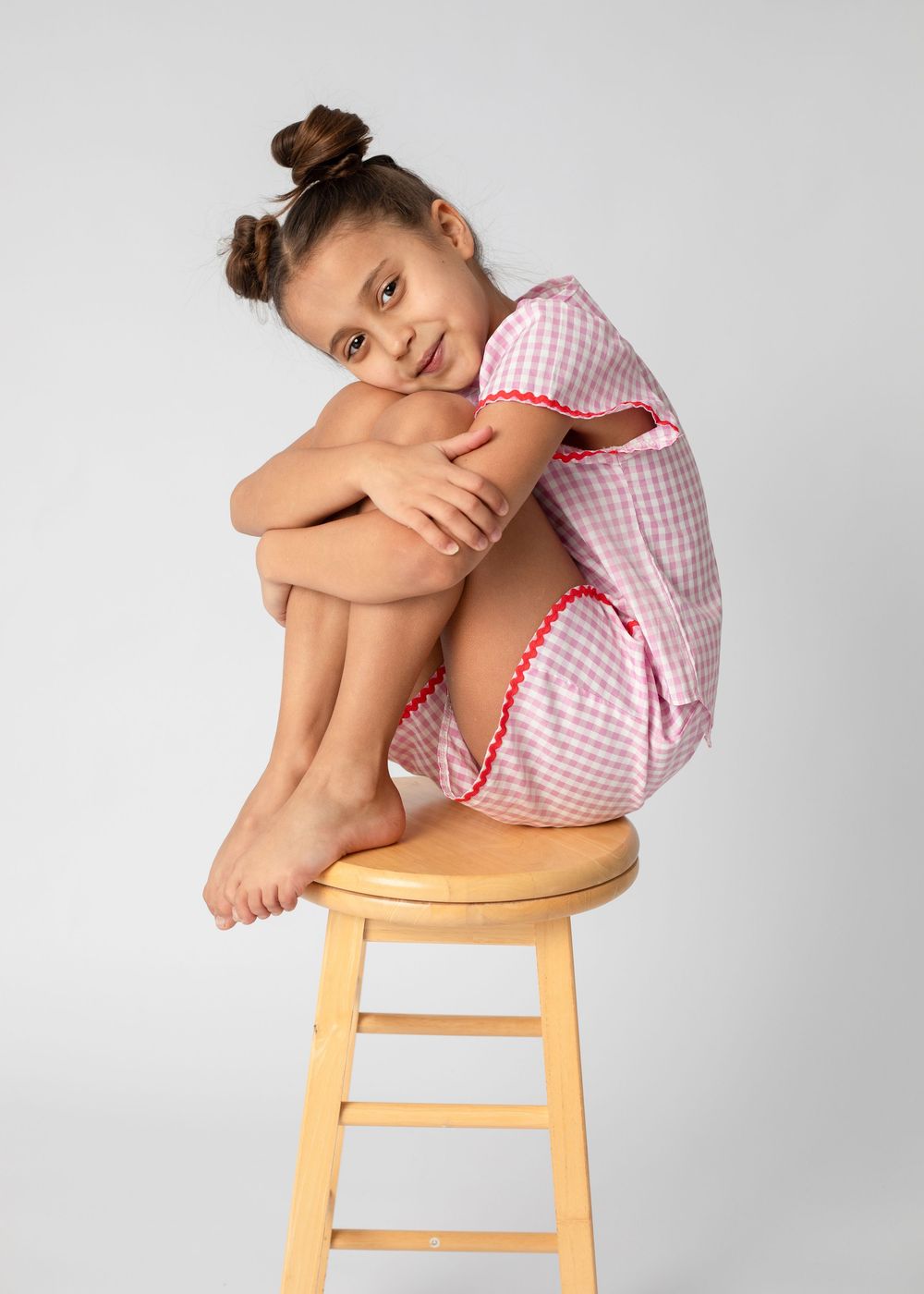 A small child in a pink gingham dress sits curled up on a wooden stool against a gray background.