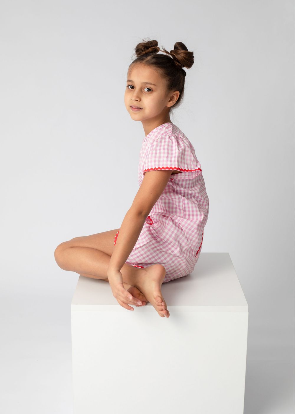 Young child in pink gingham dress sitting on white cube pedestal with hair styled in double buns.
