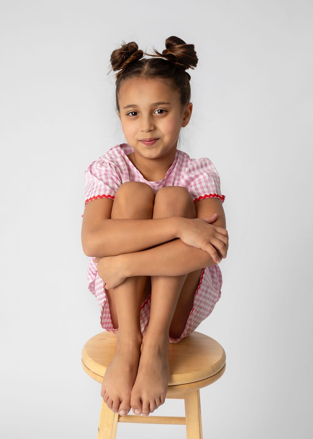 A young child in a pink gingham dress sits with knees pulled up on a wooden stool against a light background.