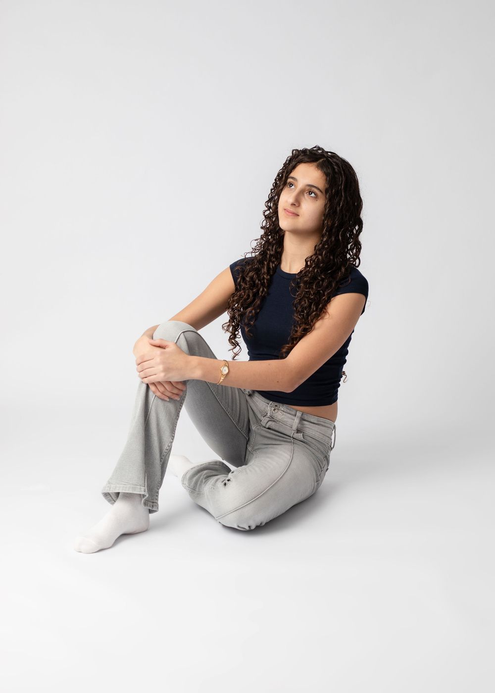 Model in a navy crop top and light gray pants sits casually on a white studio floor with long curly hair.