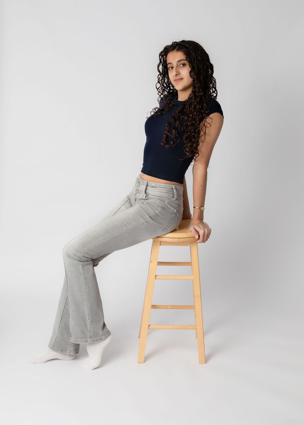 Studio portrait of someone in a navy crop top and light gray flared pants posing on a wooden stool against white background.