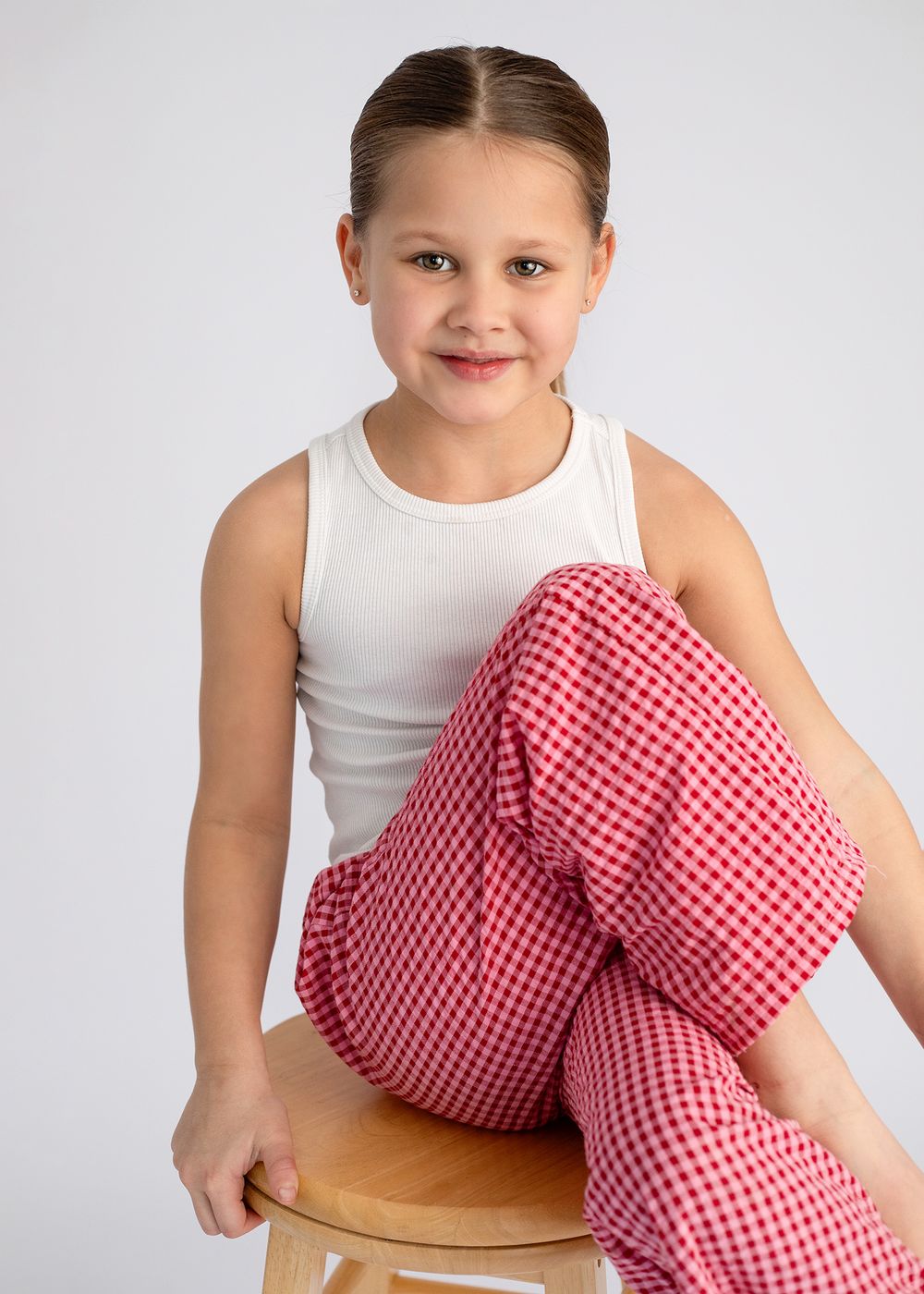 Child in white tank top and red gingham pants poses playfully on wooden stool against white background.
