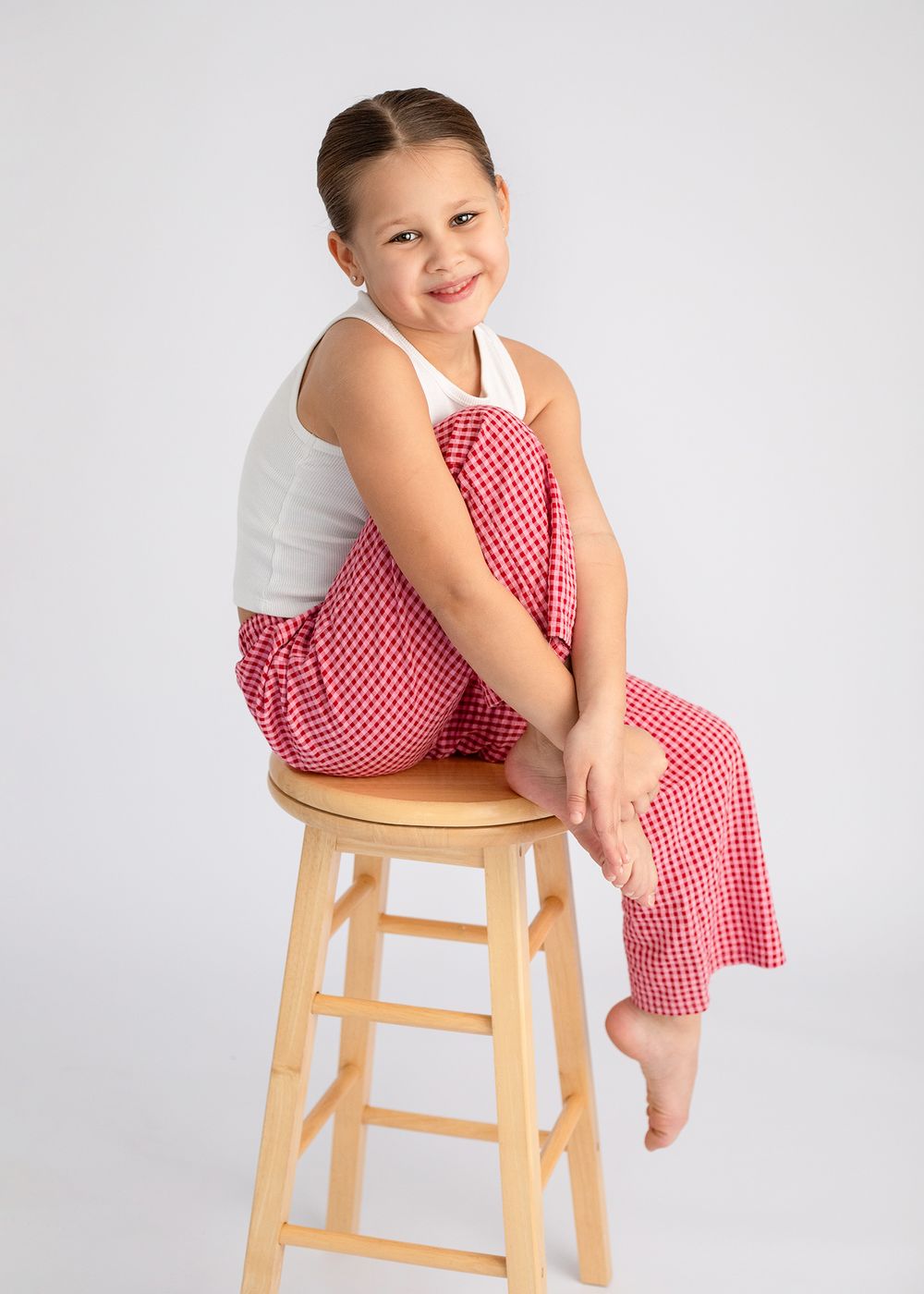 A child in a red and white polka dot dress sits playfully on a wooden stool against a light background.