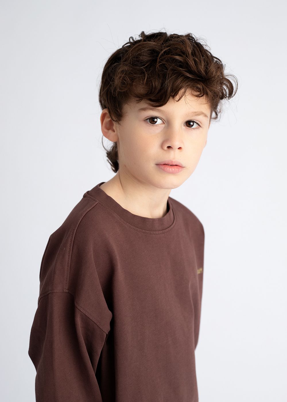 Child wearing brown sweatshirt poses against white background in studio portrait series.