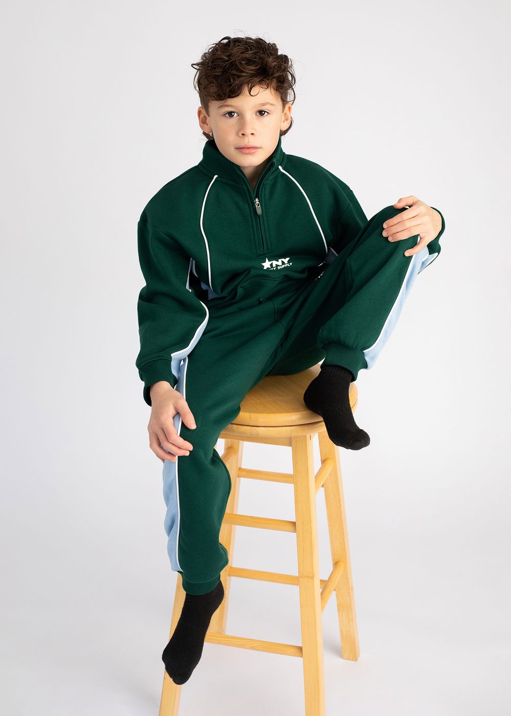 A child wearing a dark green tracksuit sits on a wooden stool against a white background.