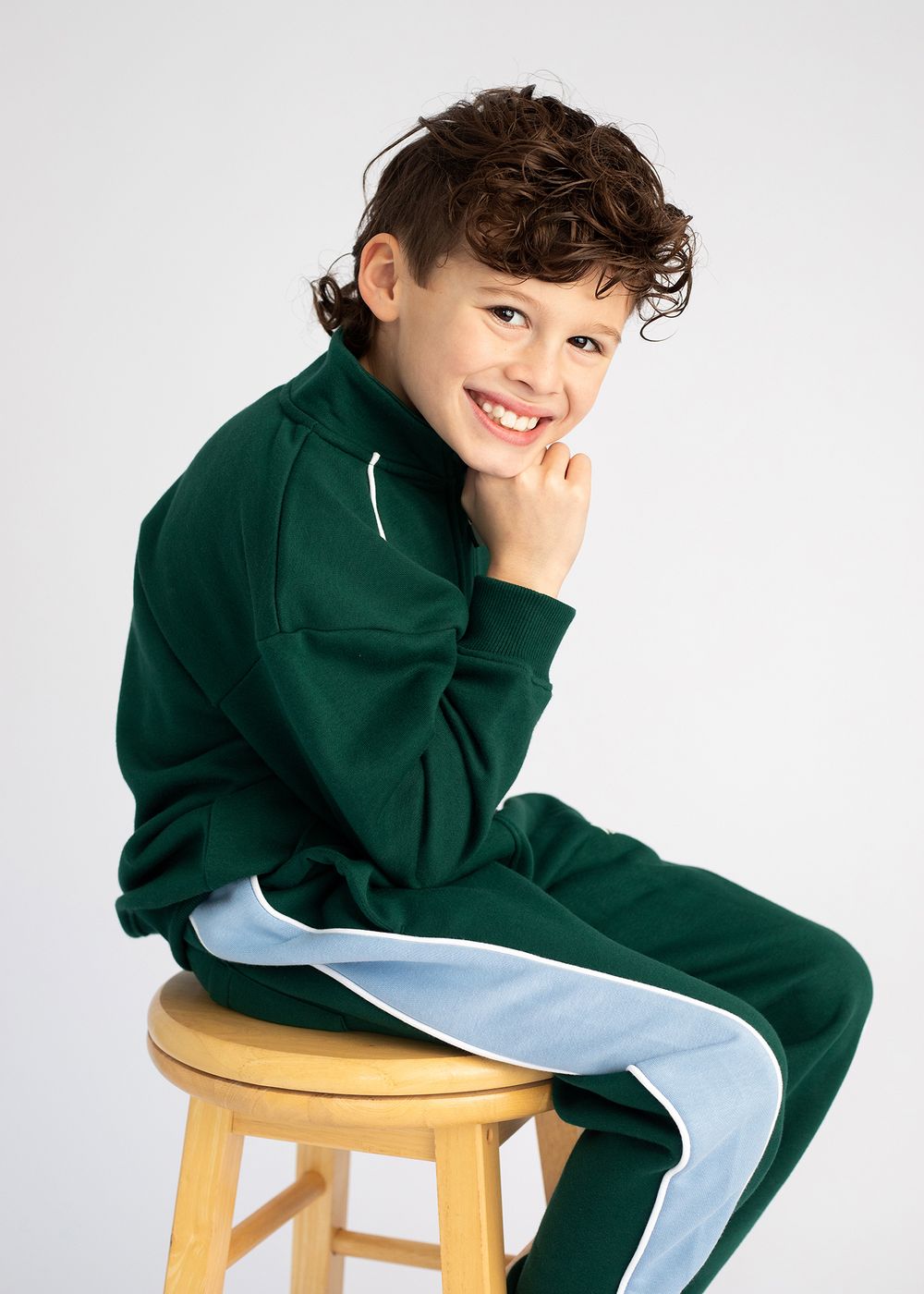 Child in dark green tracksuit sits on wooden stool against white background in four different poses.