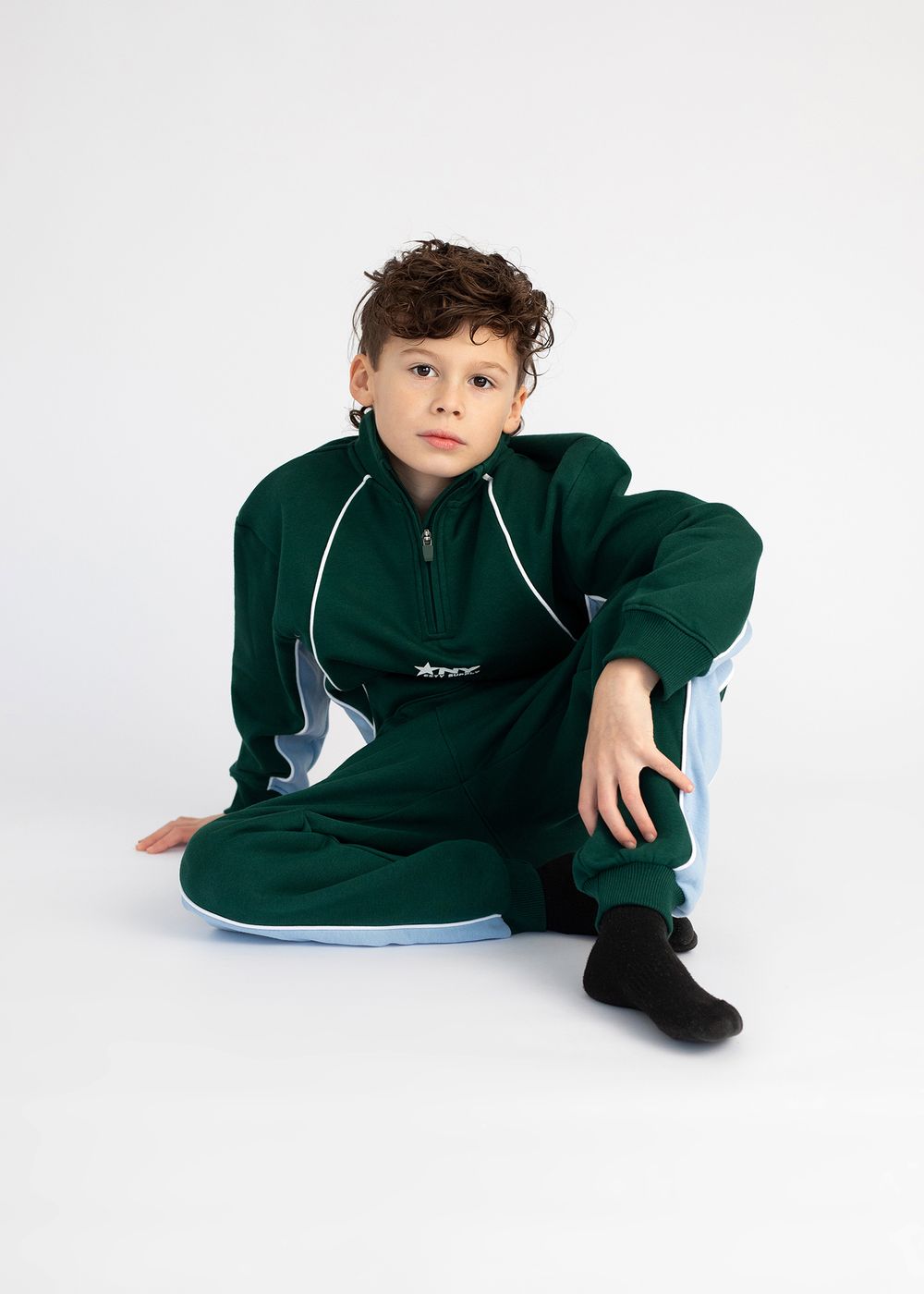 Child in dark green tracksuit lounging on white background in four different casual poses.