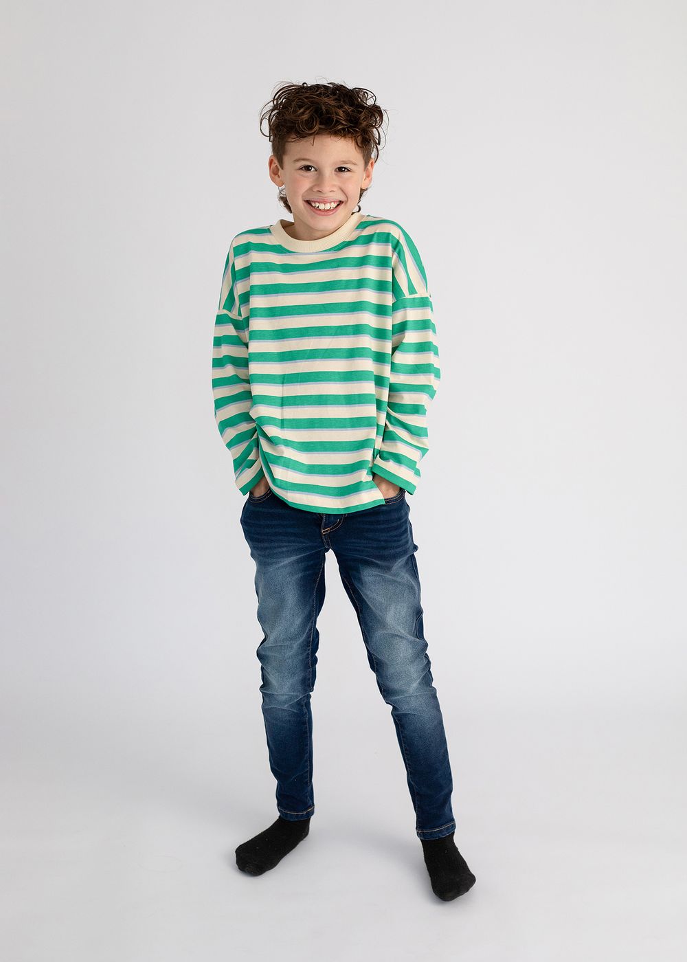 Young person wearing a green and white striped sweater and blue jeans stands casually against a light gray background.