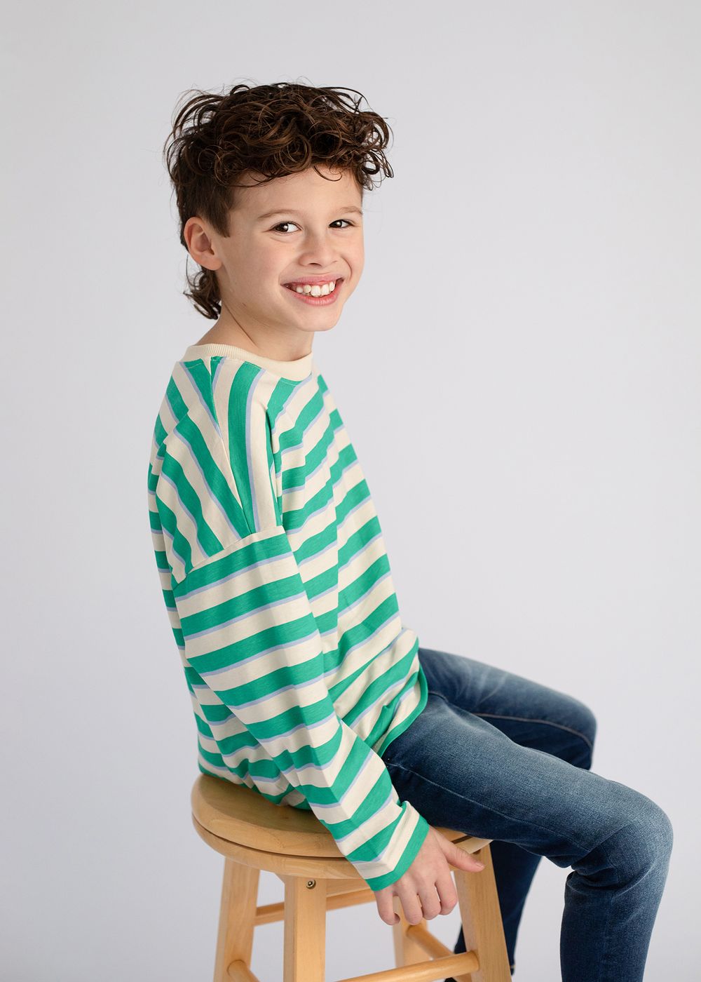 Child wearing green striped sweater and jeans sits casually on wooden stool in studio setting.