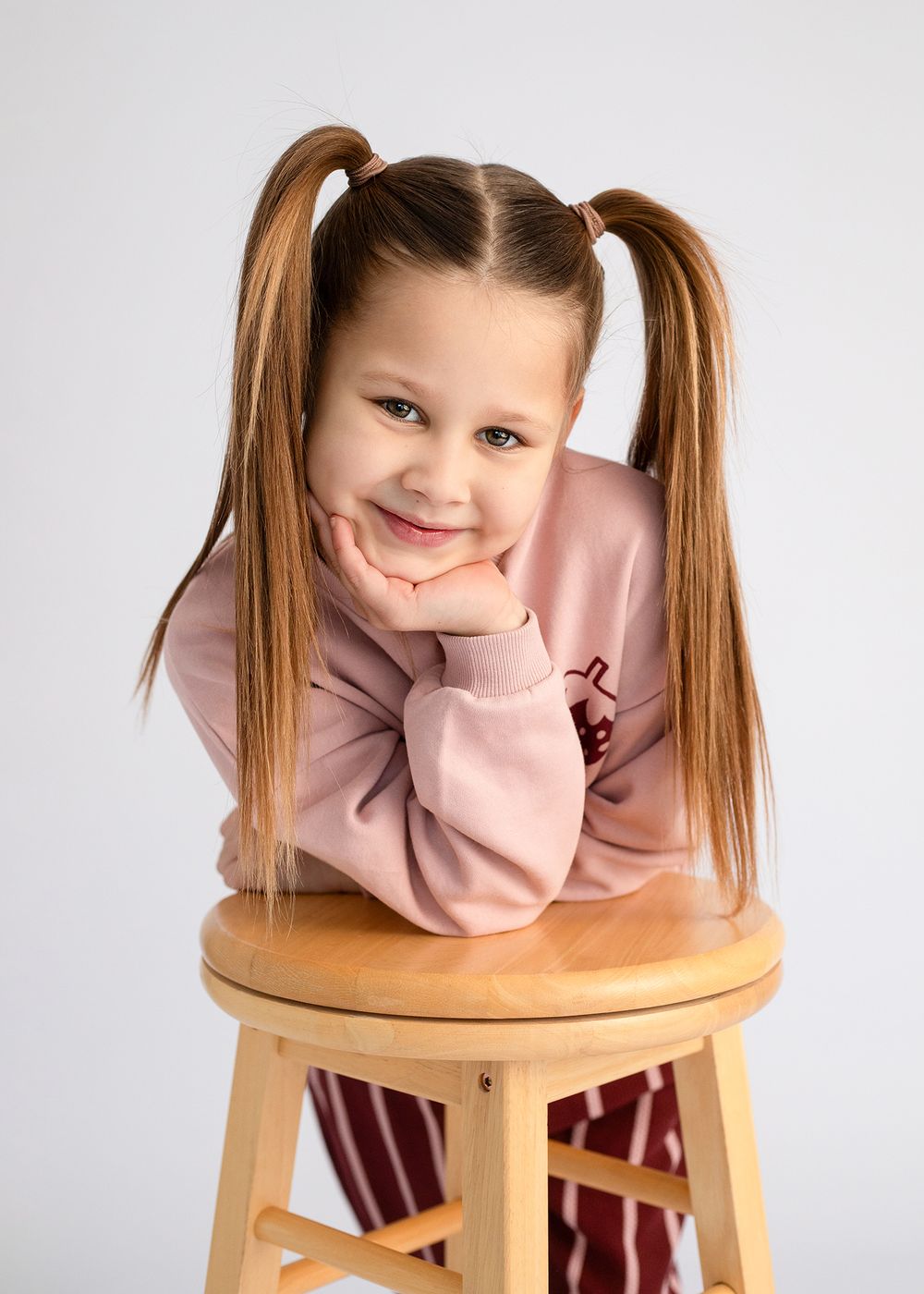 Young child in pink sweatshirt with pigtails poses with chin resting on hand while sitting on wooden stool against gray background.