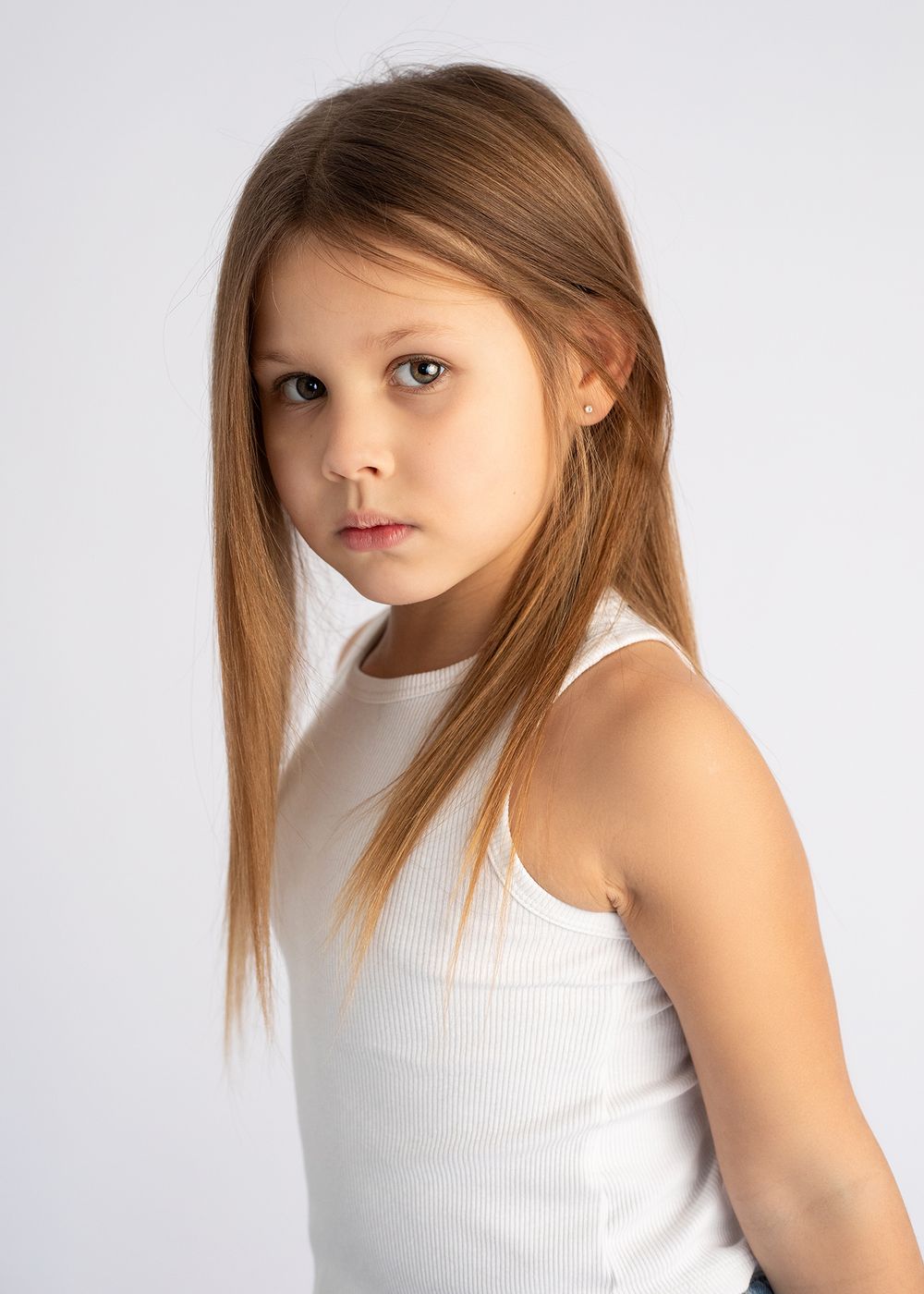 Portrait photograph of a young person with long straight brown hair wearing a white sleeveless top against a light gray background.