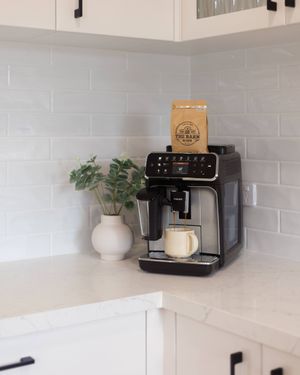 White kitchen countertop with coffee maker, potted plant, and sleek modern appliances against light tile backsplash.