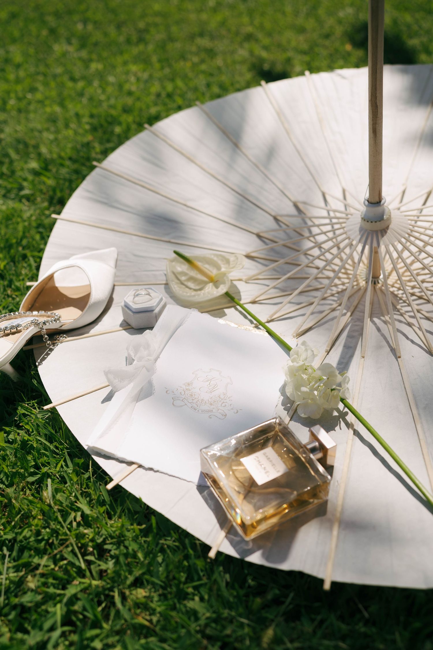 Parasol blanc sur herbe avec bouteille de parfum, fleur et verres créant un cadre extérieur élégant à la lumière du soleil.