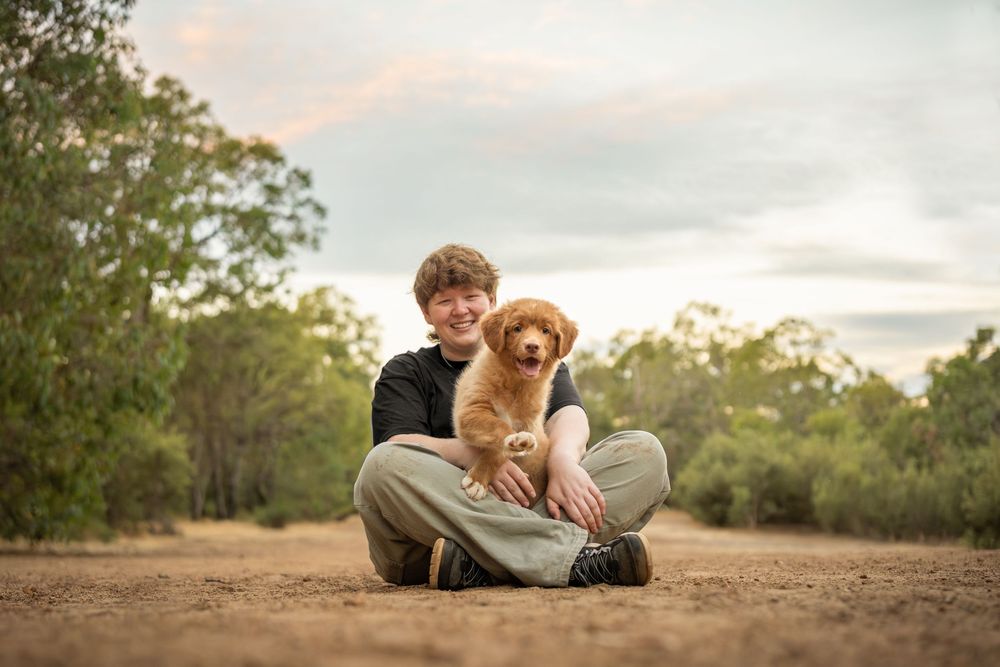 Person sitting cross-legged on dirt path holding a puppy, with trees and sunset sky behind.