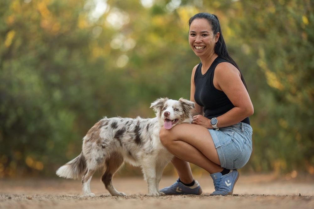 Woman smiling and crouching beside a merle Australian Shepherd dog on a dirt path surrounded by trees.