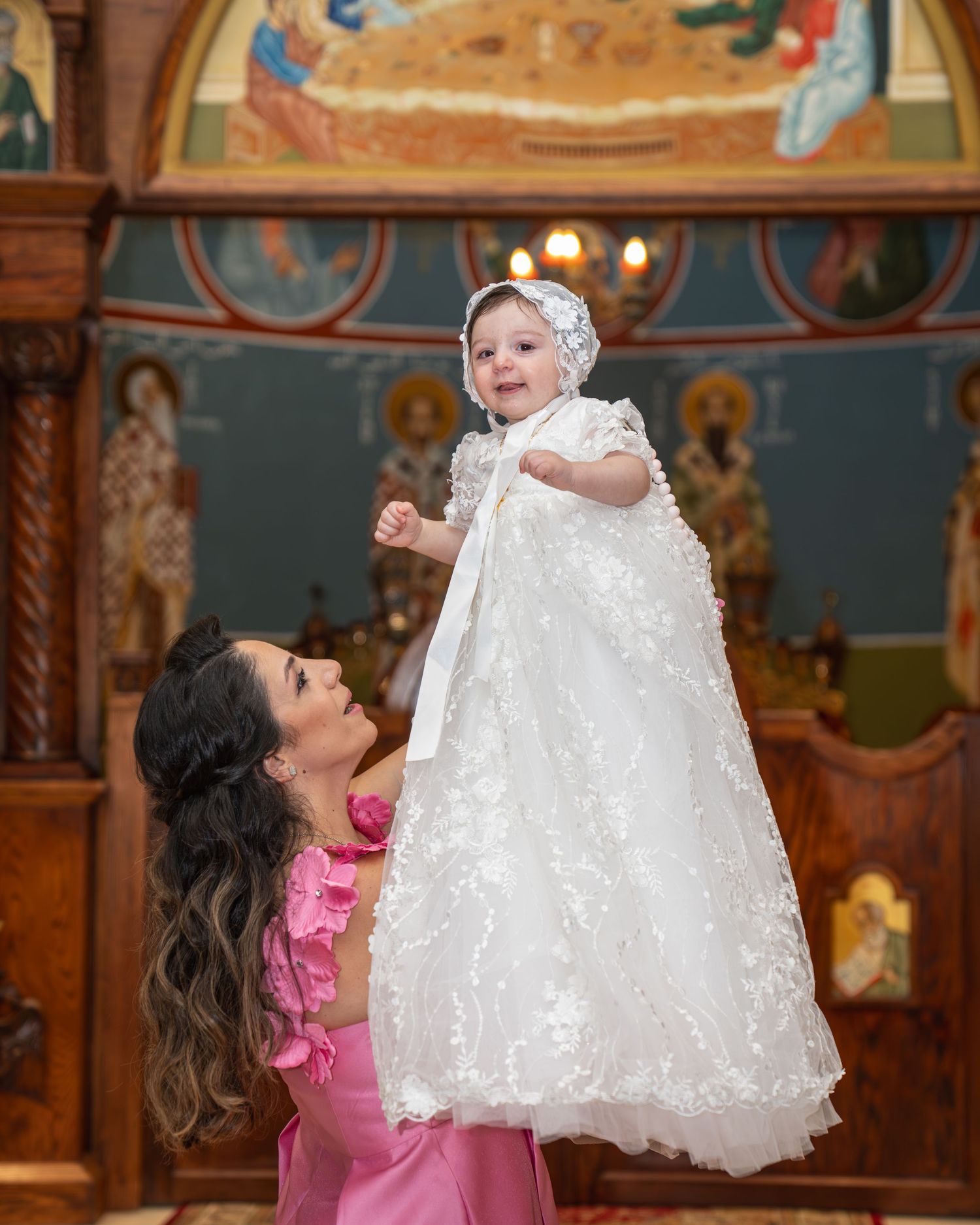 Woman in pink dress lifting smiling baby in white lace christening gown inside ornate Orthodox church.