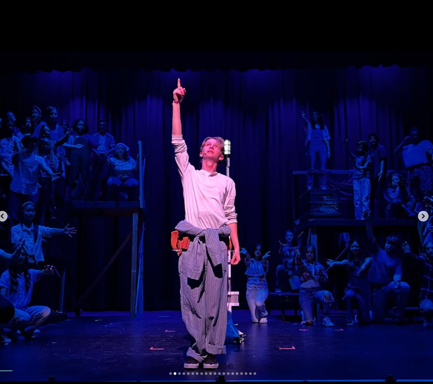 A young male performer points upward dramatically on a blue-lit stage during a theatrical production.