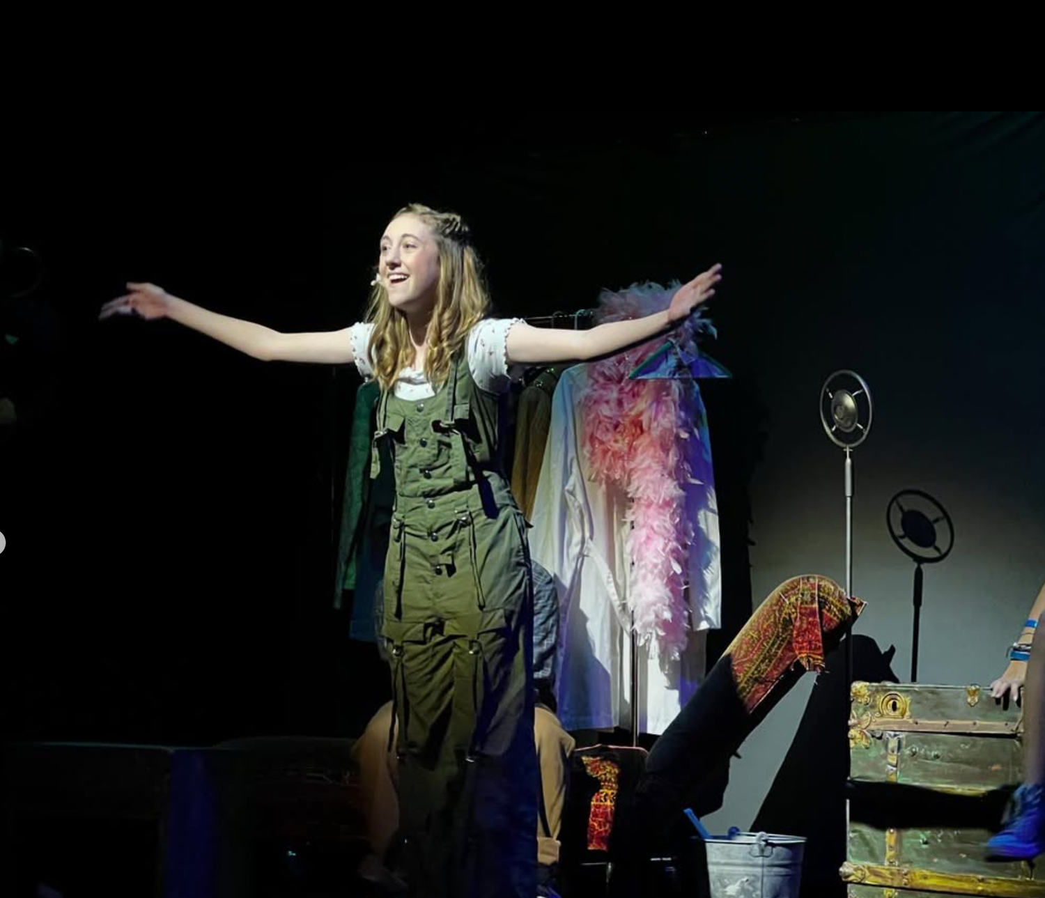 A young girl in overalls performs with outstretched arms on a darkened theatrical stage.