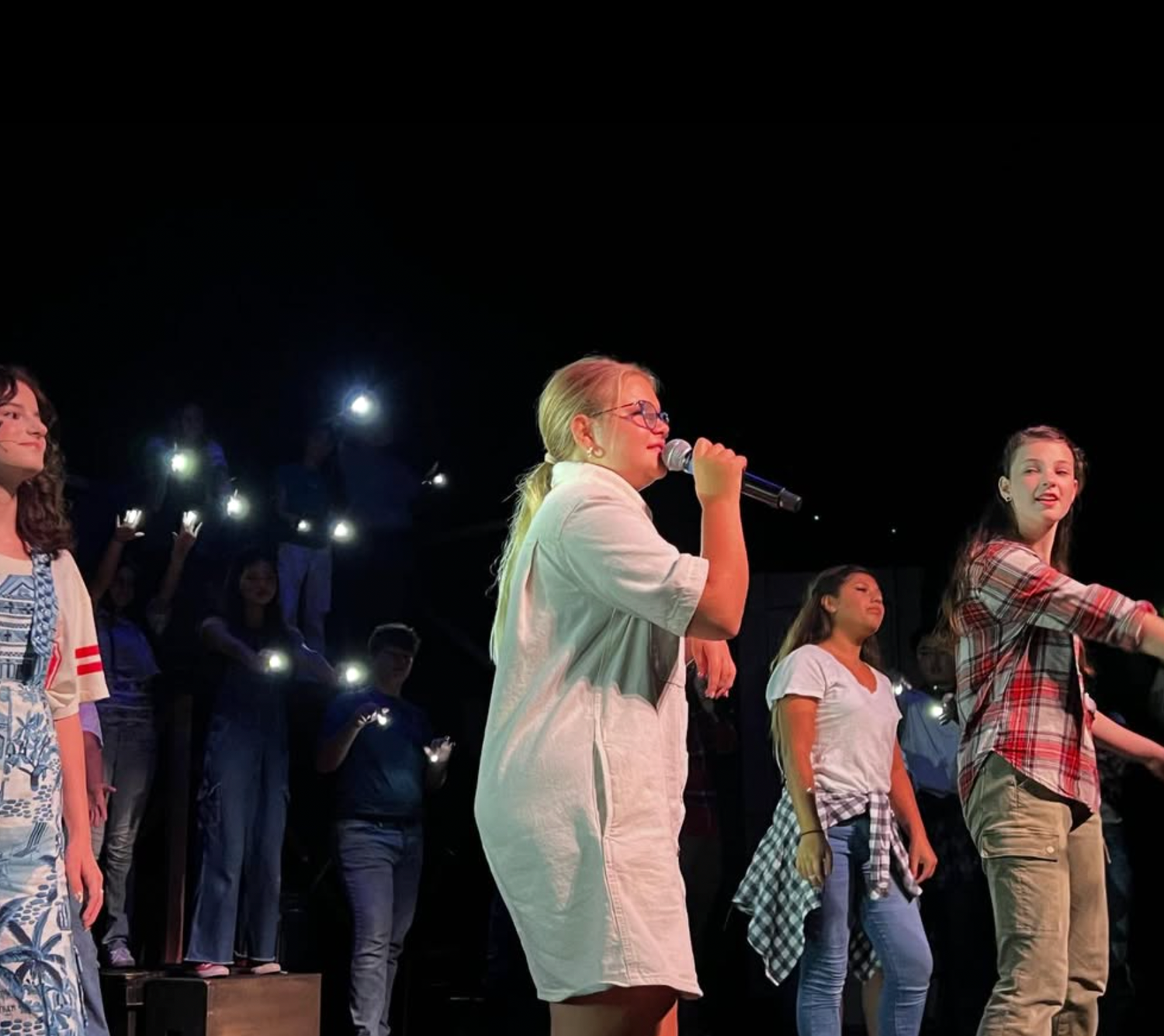 Students perform on stage holding microphones with audience members lighting phones in the background.