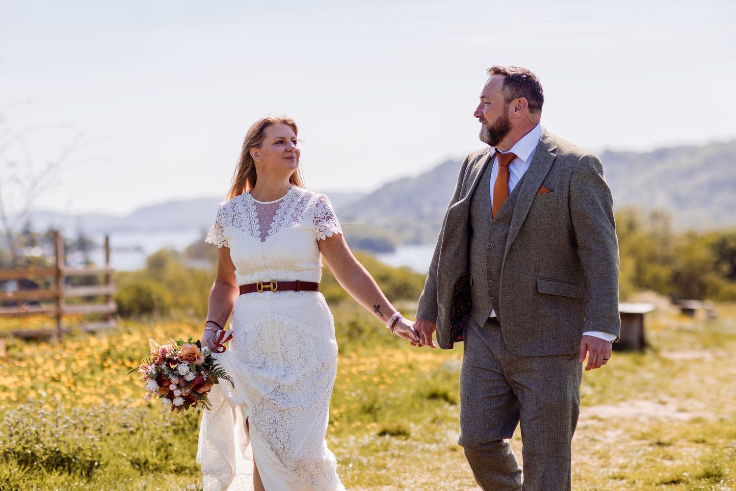 Bride and groom walk hand-in-hand outdoors on sunny day, she holds bouquet in white lace dress, he wears brown tweed suit.