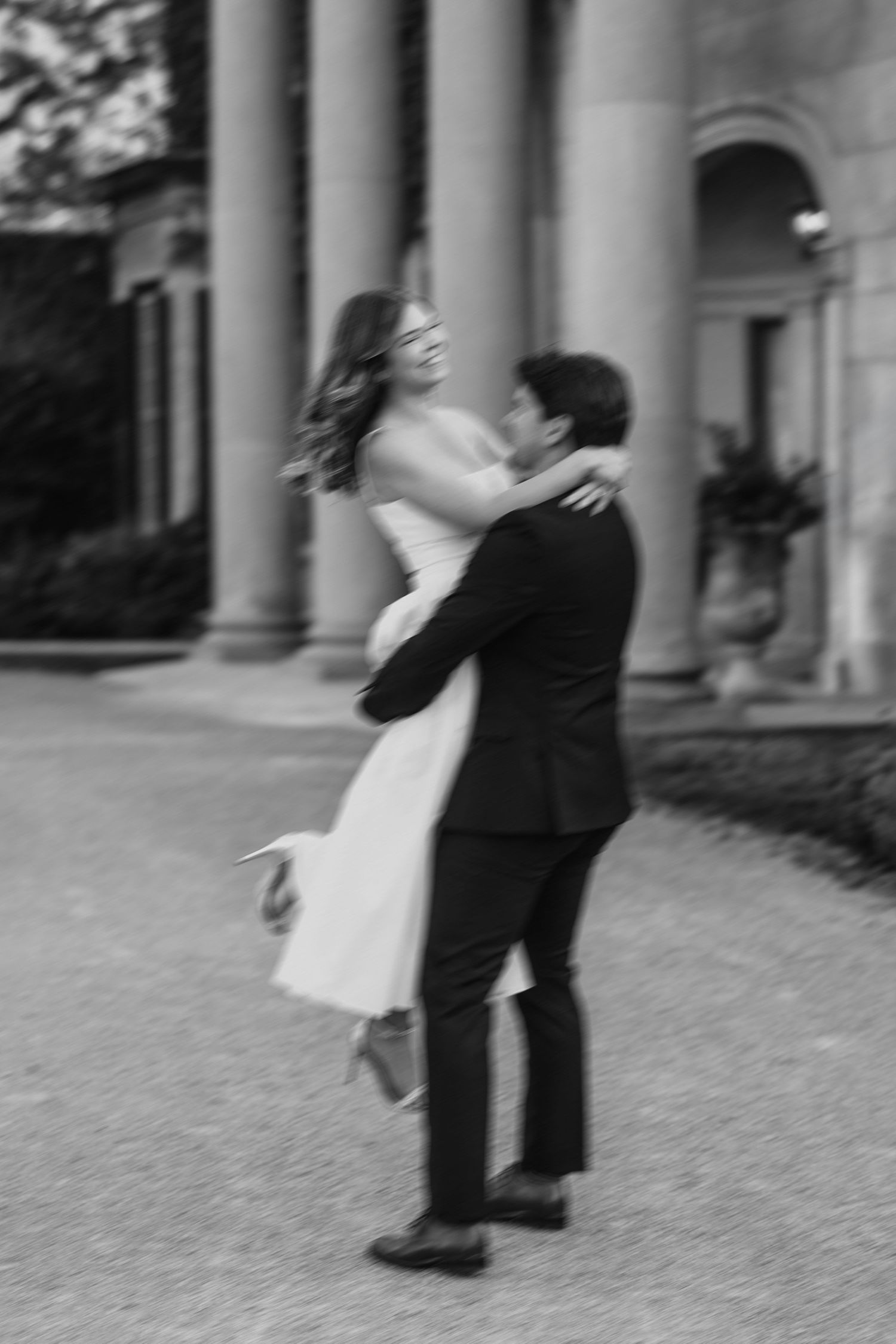 Black and white photo of a groom lifting his bride outdoors near elegant columns.