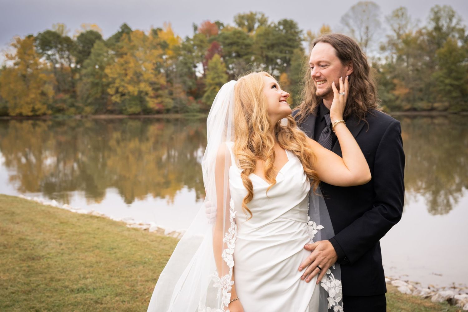 Bride and groom share a romantic moment by a lake surrounded by autumn foliage, bride in white gown with veil.