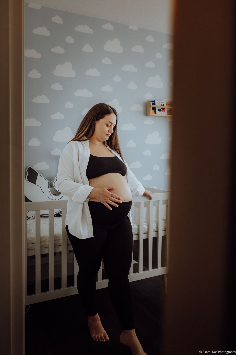 Pregnant woman in black outfit cradling her baby bump in a nursery with cloud-patterned wallpaper and white crib.