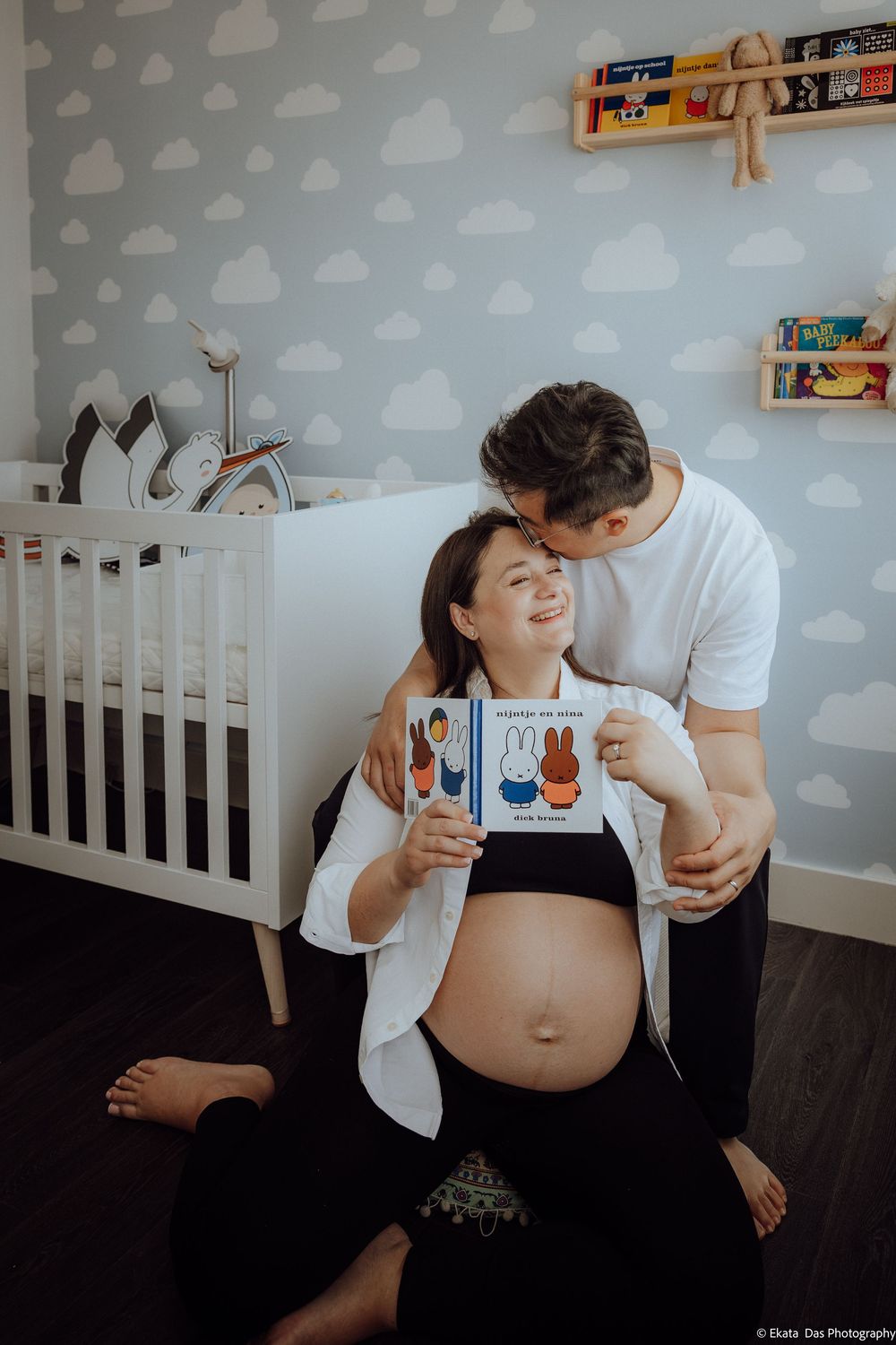 Couple in nursery, pregnant woman holding Miffy book, partner kissing her head, white crib and cloud wallpaper background.