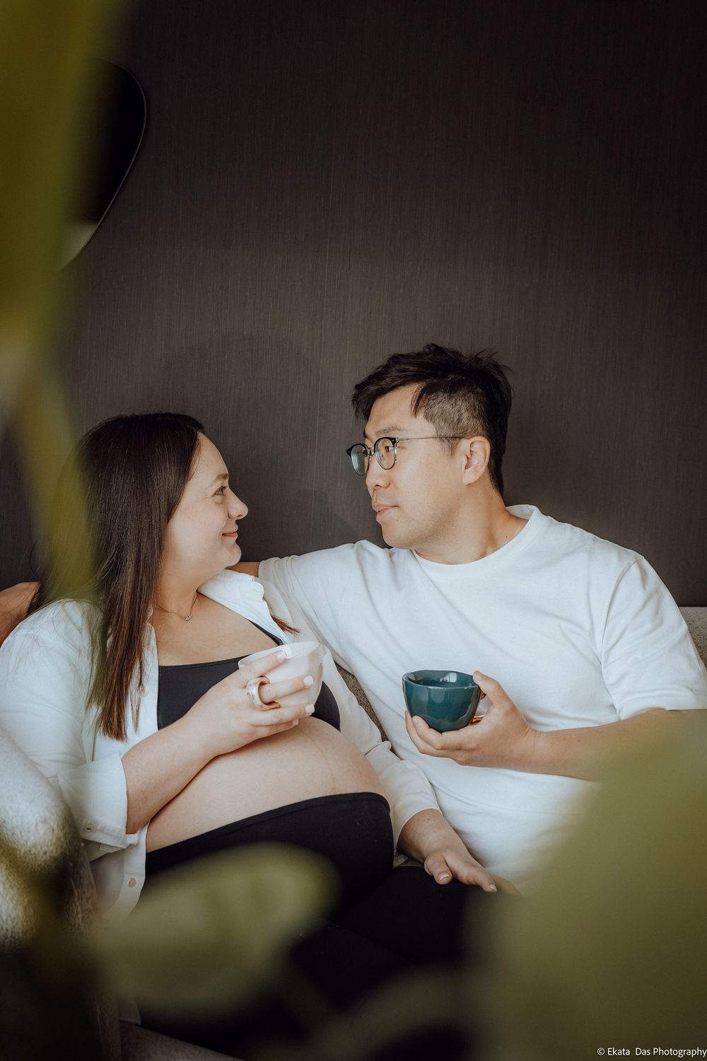 Pregnant couple sitting on couch sharing coffee, smiling at each other in a cozy indoor setting.