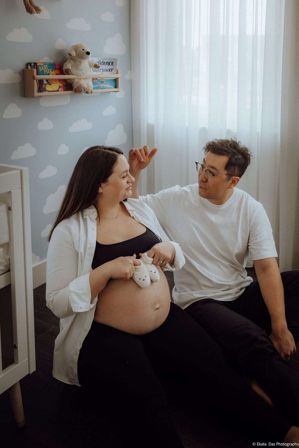 Pregnant woman holding baby shoes on her belly sitting with her partner in a decorated nursery room.