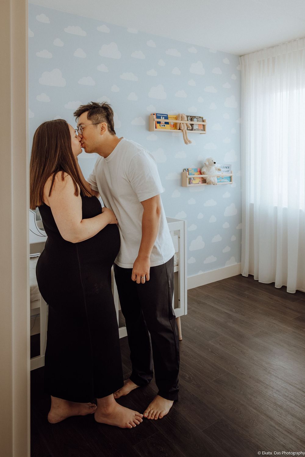 Couple sharing a tender kiss in a baby nursery with cloud wallpaper, woman in black dress showing baby bump.