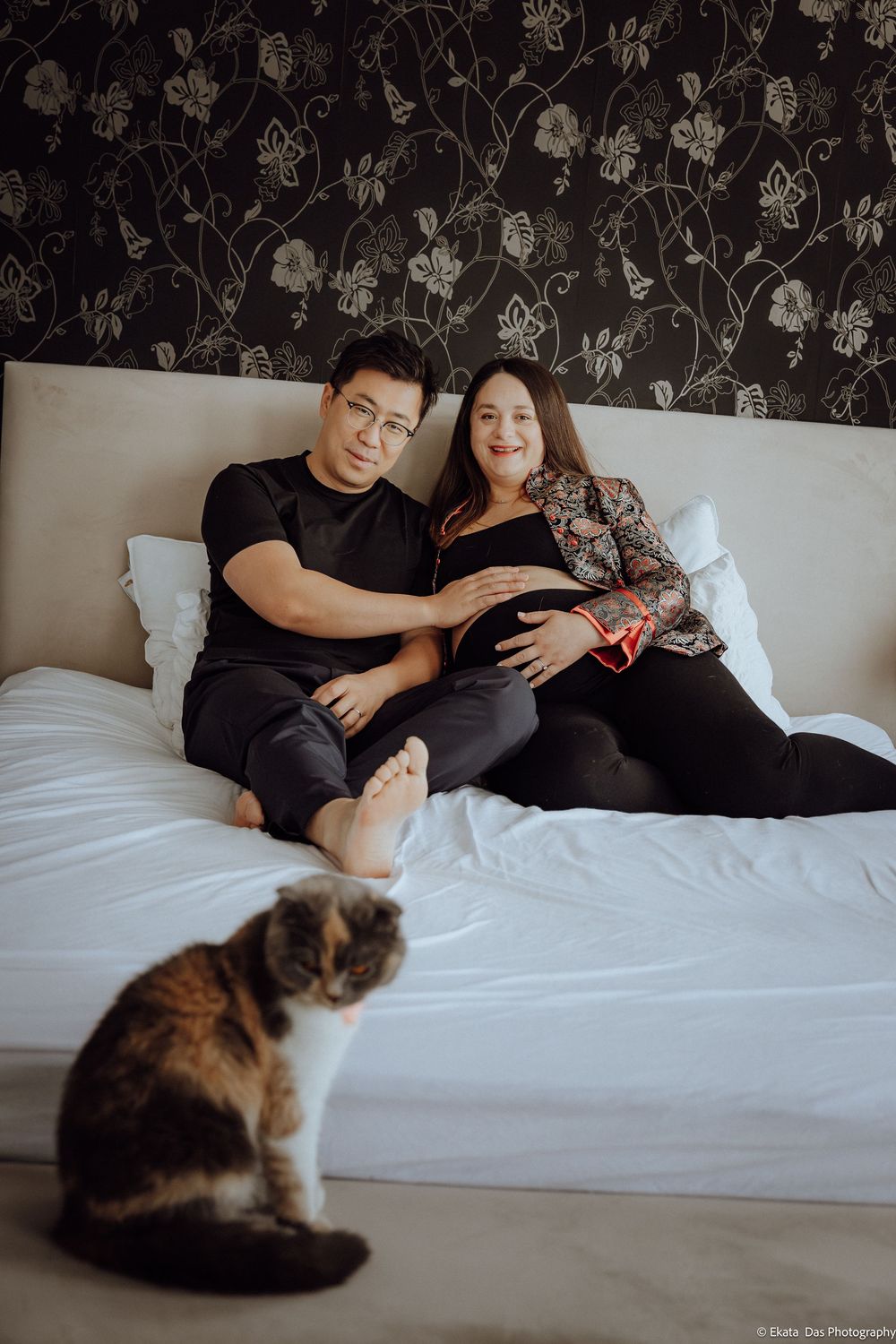 Couple sitting on bed with cat in foreground, woman showing baby bump, dark floral wallpaper backdrop.
