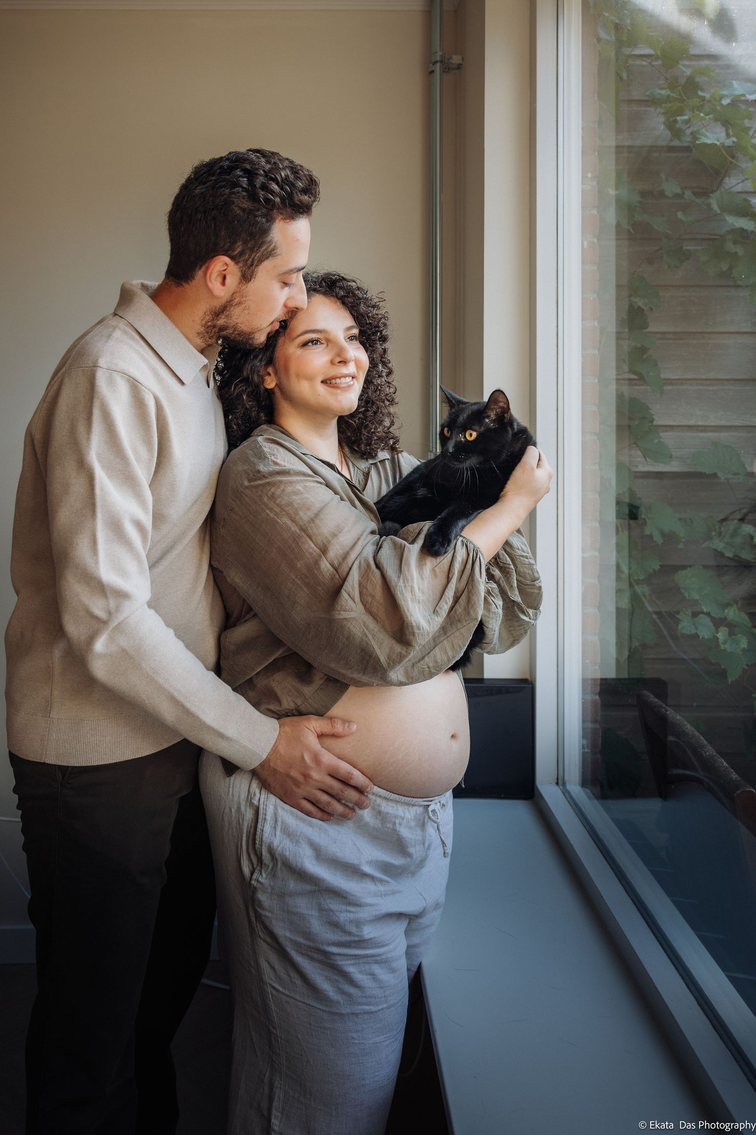 Expectant couple embracing by a sunlit window as the smiling pregnant woman holds a black cat in a warm maternity photo.